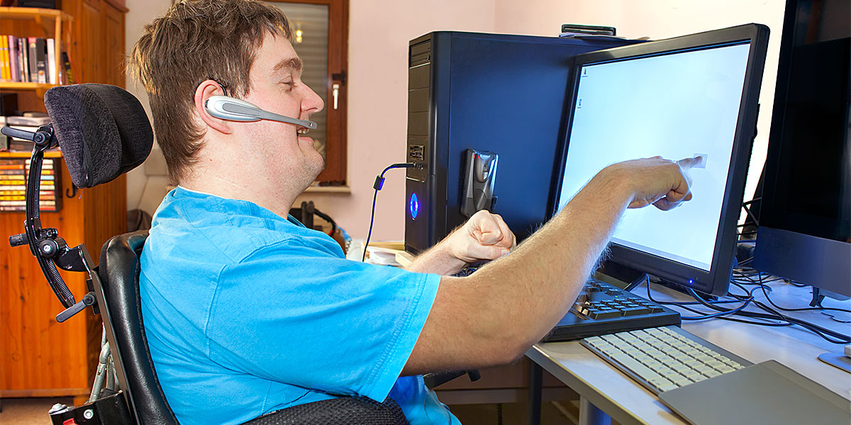 Spastic young man with infantile cerebral palsy caused by a complicated birth sitting in a multifunctional wheelchair using a computer with a wireless headset reaching out to touch the touch screen