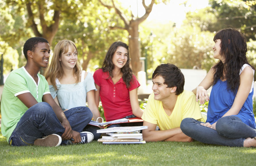 Five students sitting on the lawn and smiling with books open